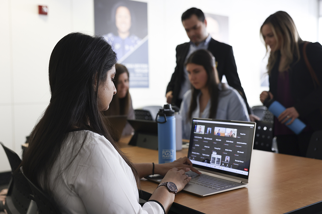 A GWSB student works on a laptop in duques Hall while other students chat with a professor in the background