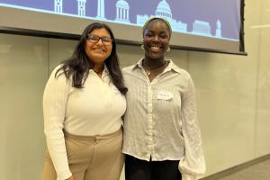 Two people stand side by side smiling. A projection can be seen on a wall behind them.